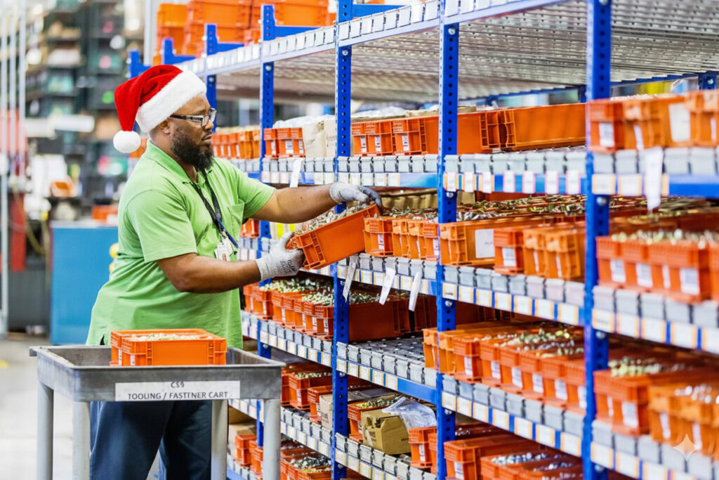 A worker in a Santa hat organizes orange storage bins on shelves in a warehouse. The scene conveys a festive spirit and industrious atmosphere. FlexMation.
