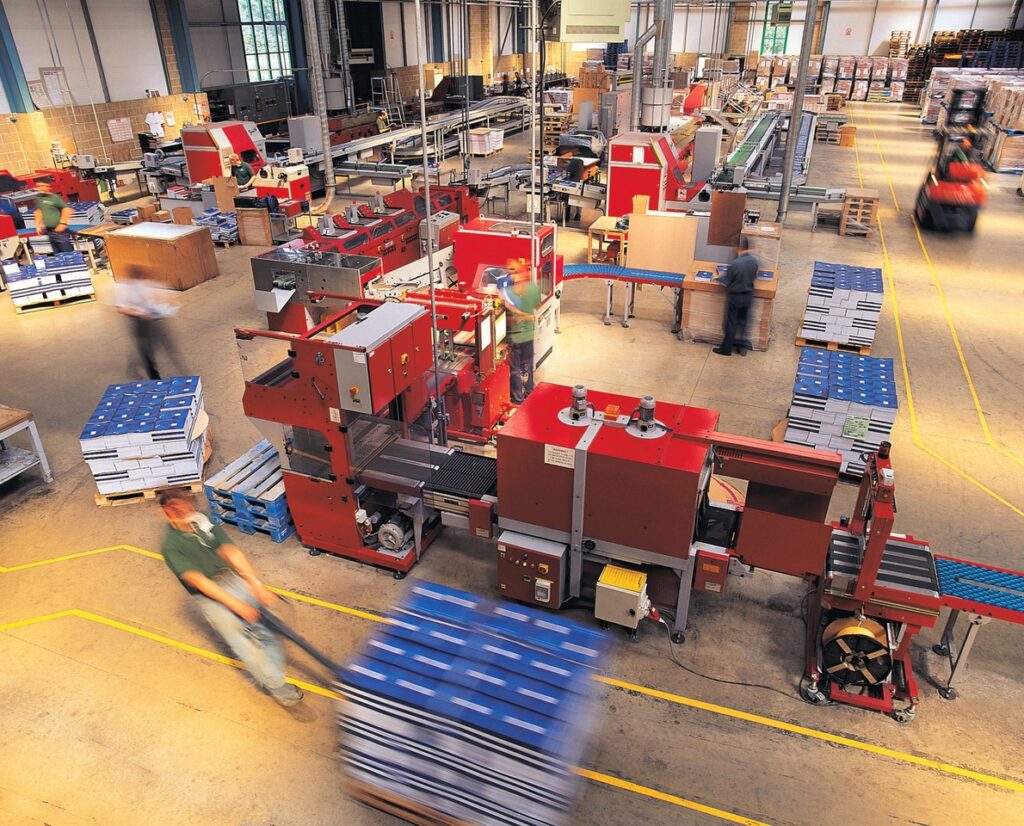 A busy packaging factory with red machinery, stacks of blue and white pallets, and workers in motion, conveying efficiency and industriousness. FlexMation