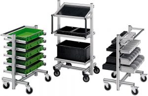 Three mobile industrial workstations made from aluminum framing and mounted on caster wheels. The left workstation features multiple pull-out shelves holding stacked green plastic storage bins. The center workstation includes a slanted work surface with shelves supporting black plastic totes. The right workstation has several angled pull-out trays containing small black and gray parts bins for organized component storage. All three units are displayed against a light gray background. FlexMation.